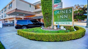 A Dunes Inn sign with a green arrow pointing left for entry, a black SUV parked in front, and a building with a modern design and a tree in the foreground.