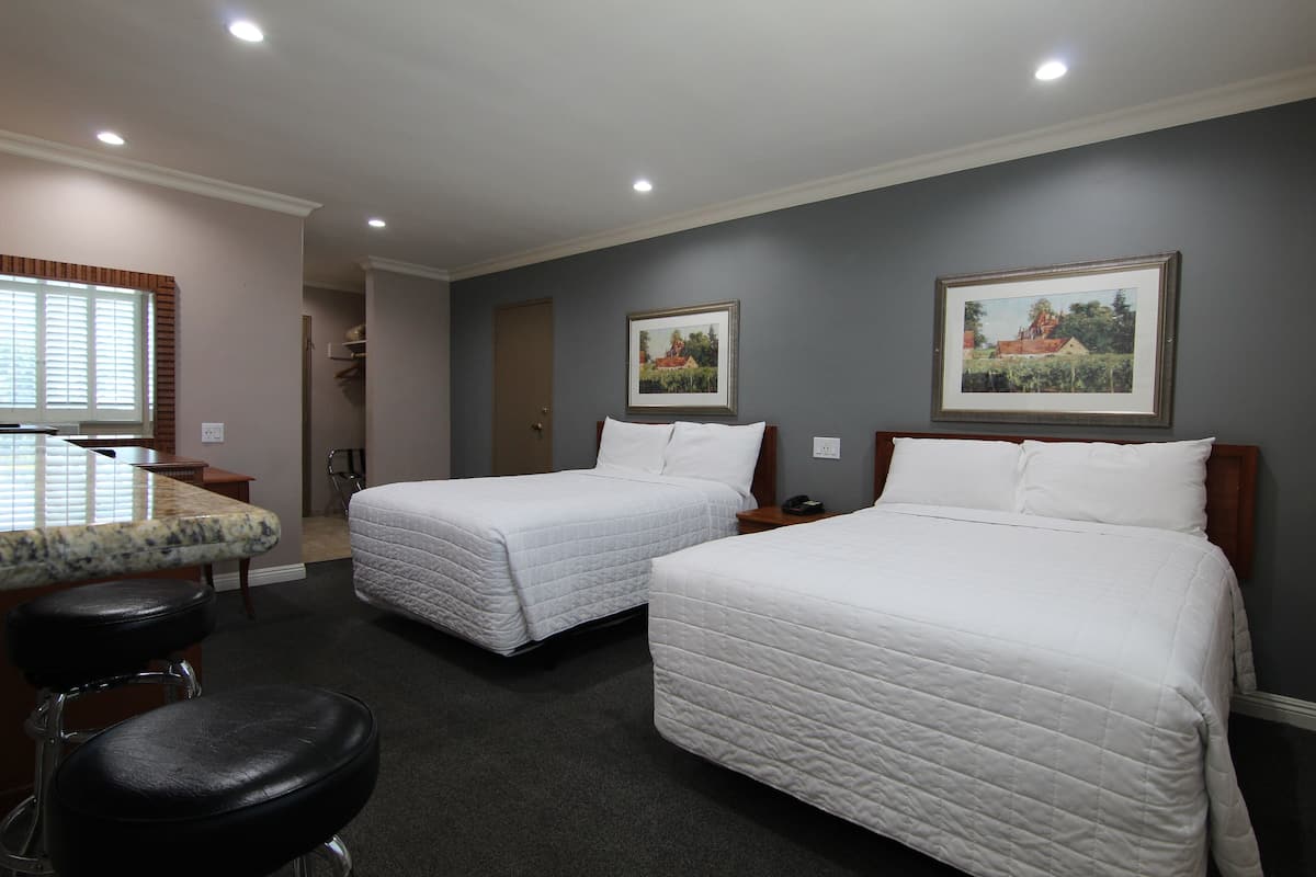 A hotel room with two beds, a small kitchenette, a bar stool, and two framed pictures on the wall.