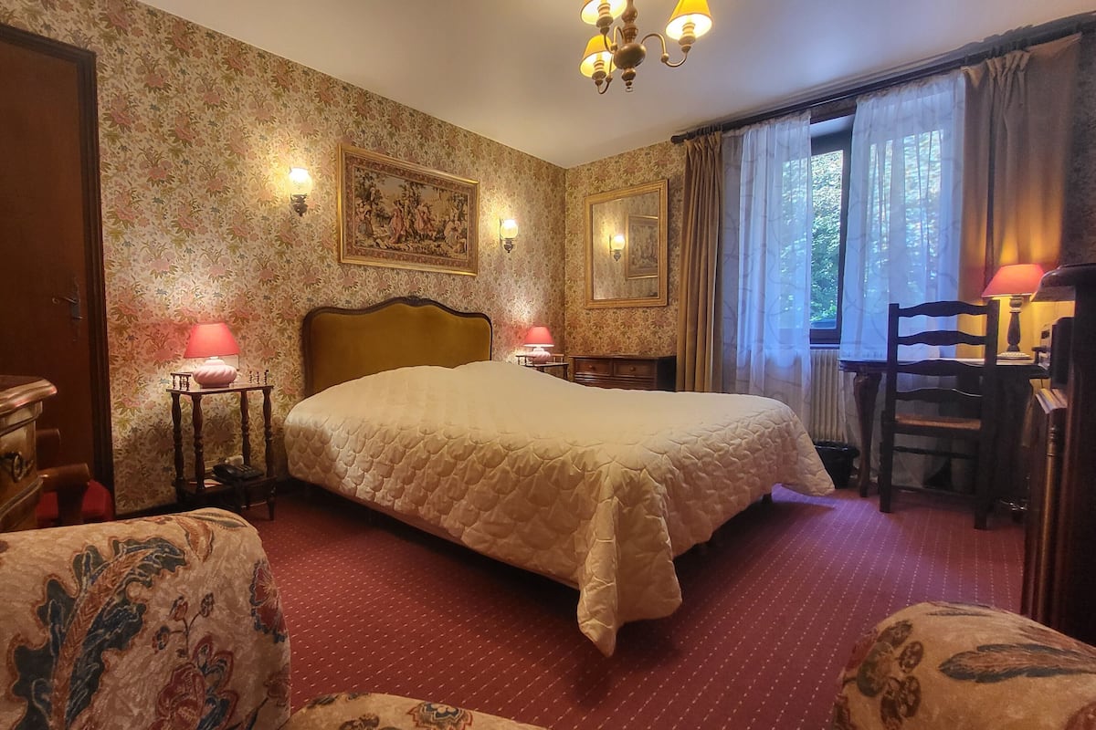 A hotel room with a bed, two bedside tables with lamps, a chair, a desk, a mirror, and a window with curtains.