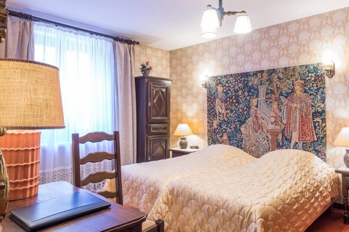 A hotel room with a bed, a desk, a chair, a lamp, a cabinet, and a tapestry on the wall.