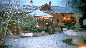 Un patio cubierto de nieve con un edificio que tiene tejado de baldosas y luces colgantes. Hay plantas en macetas y una fuente de piedra.