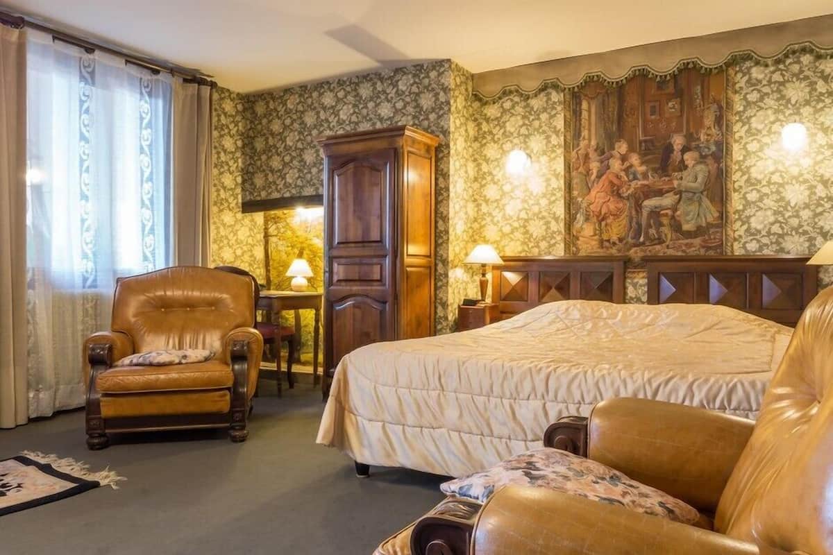 A room with a large bed, a wooden wardrobe, a leather armchair, and a table with a lamp.