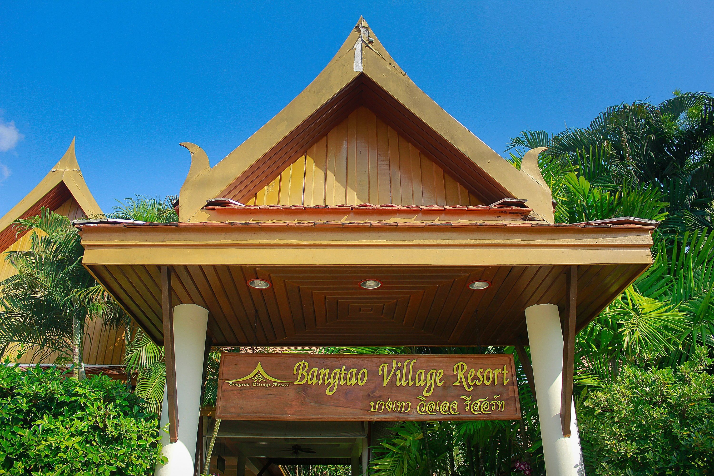 Een resort ingang met een traditionele houten constructie en een bord waarop staat 'Bangtao Village Resort'.