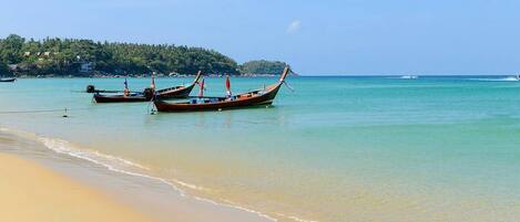 A beach with clear turquoise water and a sandy shore. There are traditional long-tail boats moored near the water's edge.