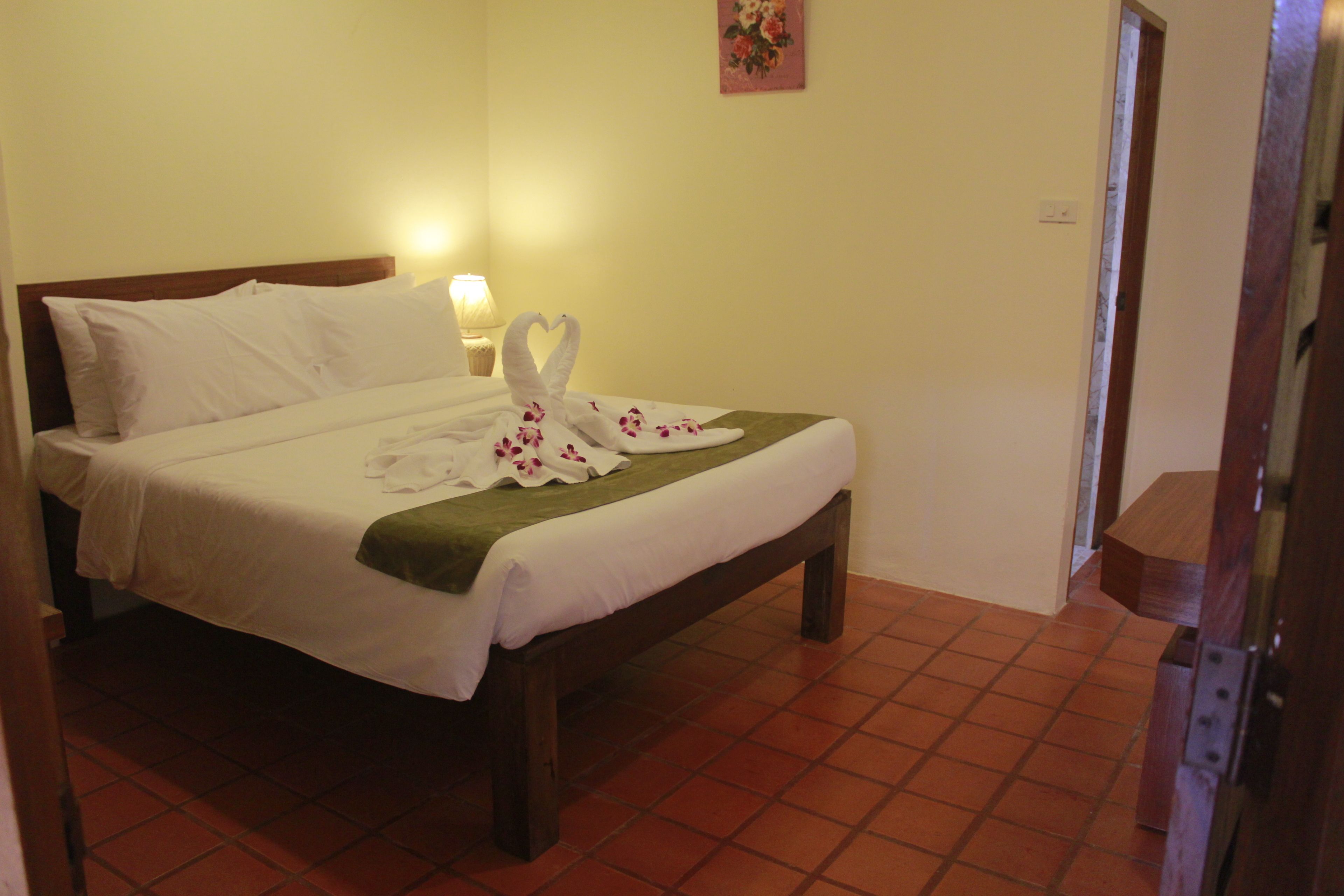 A neatly made bed with white linens and a green throw blanket, a wall-mounted lamp, and a floral painting on the wall.