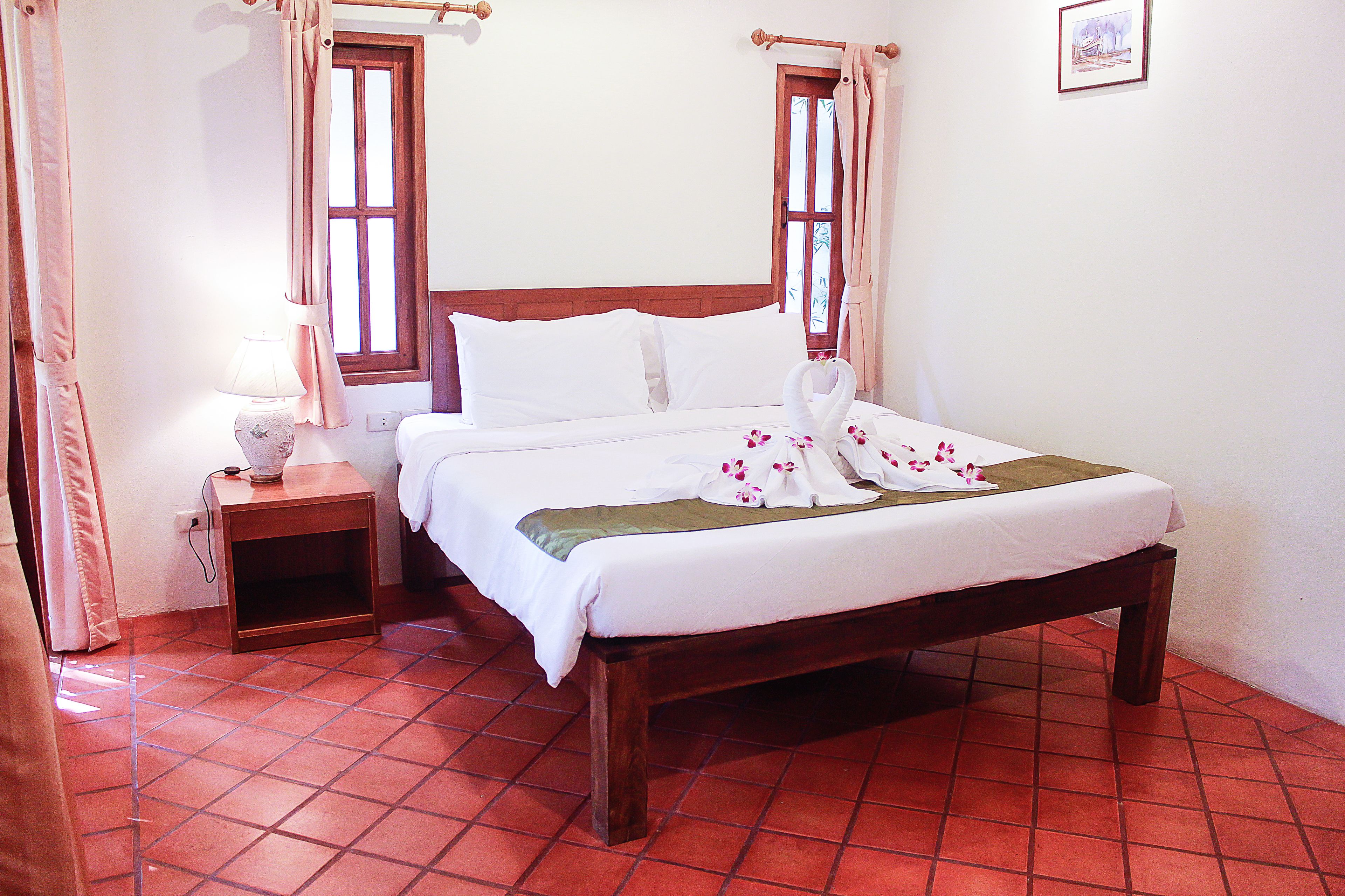 Una cama bien hecha con sábanas blancas y un plaid verde, una mesita de noche de madera con una lámpara, y una ventana con cortinas.