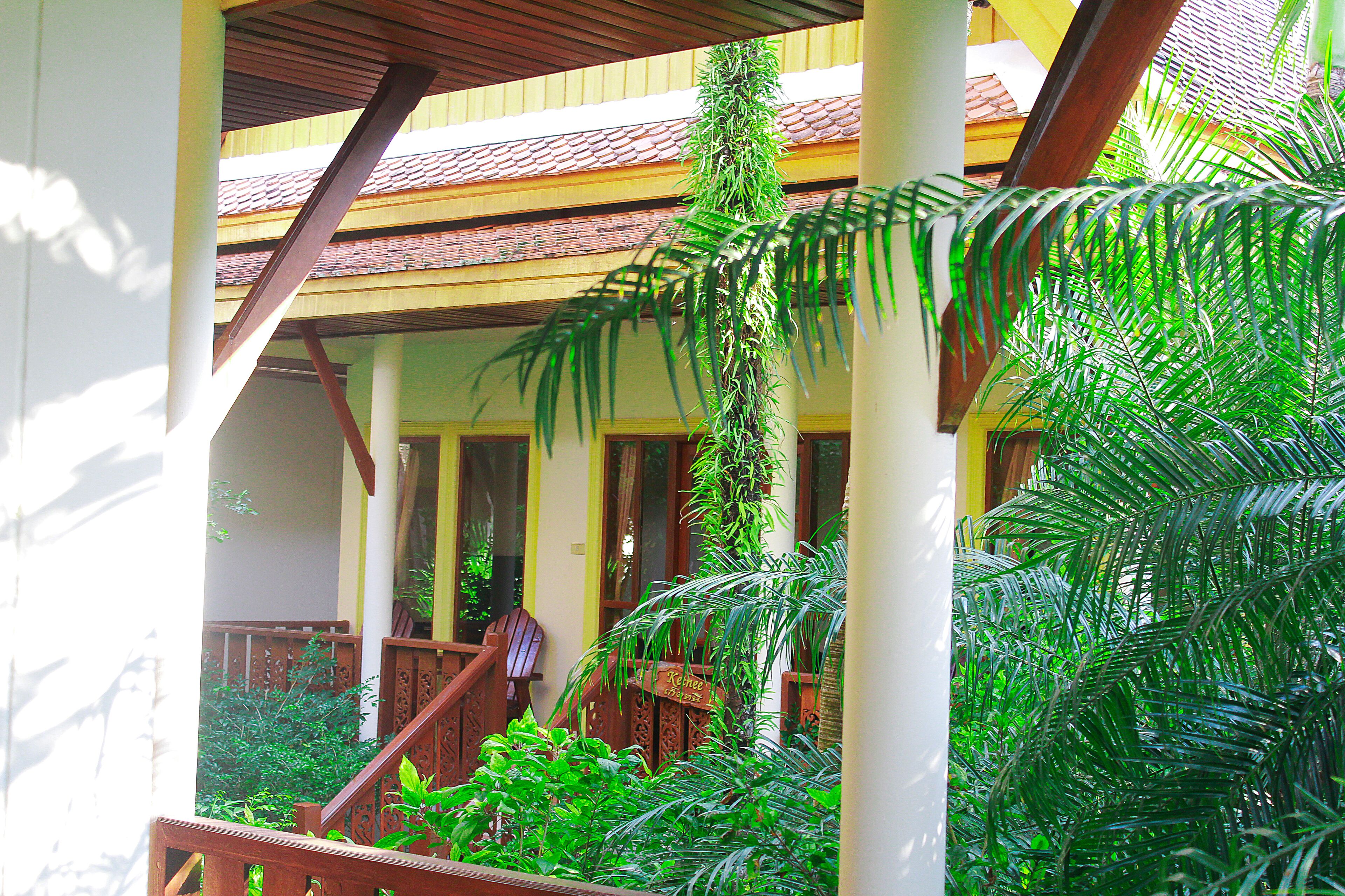 A covered outdoor area with wooden railings, potted plants, and a staircase leading to a building with a tiled roof.