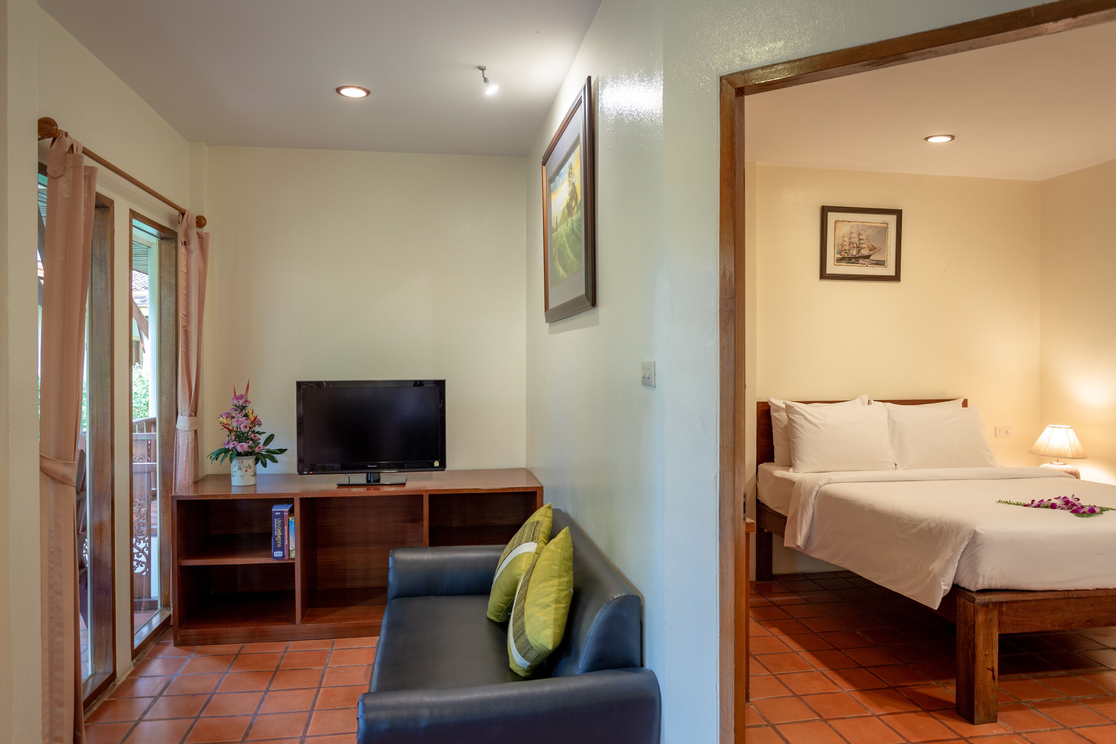 A room with a bed, a TV on a wooden stand, a sofa with cushions, and a small table with a flower vase.