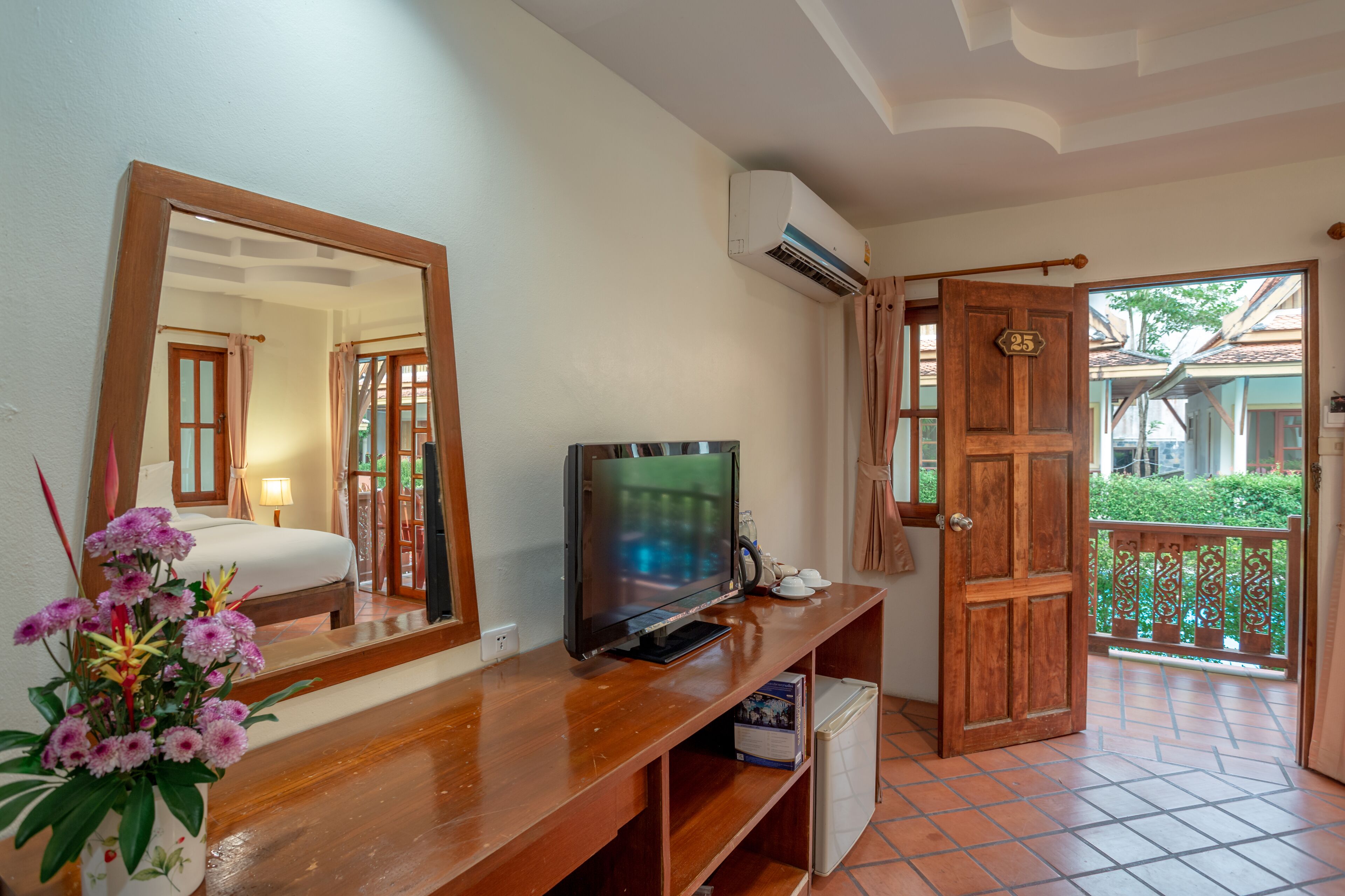 A hotel room with a wooden console table, a television, a mirror, a potted plant, and a door leading to a balcony with a view of greenery.