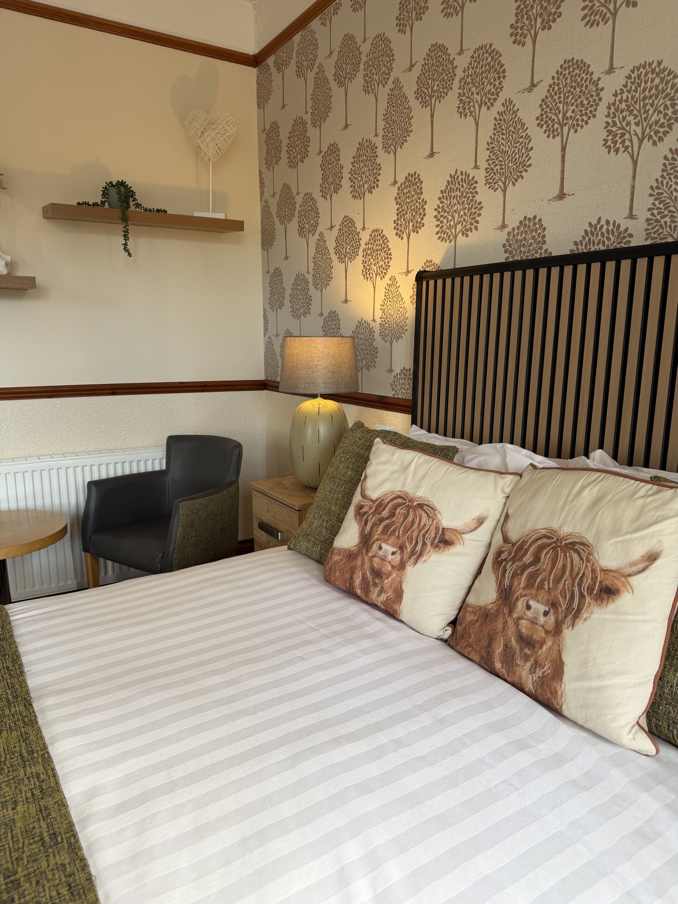 A hotel room with a bed, a chair, a table, a lamp, and a wall with tree patterns.