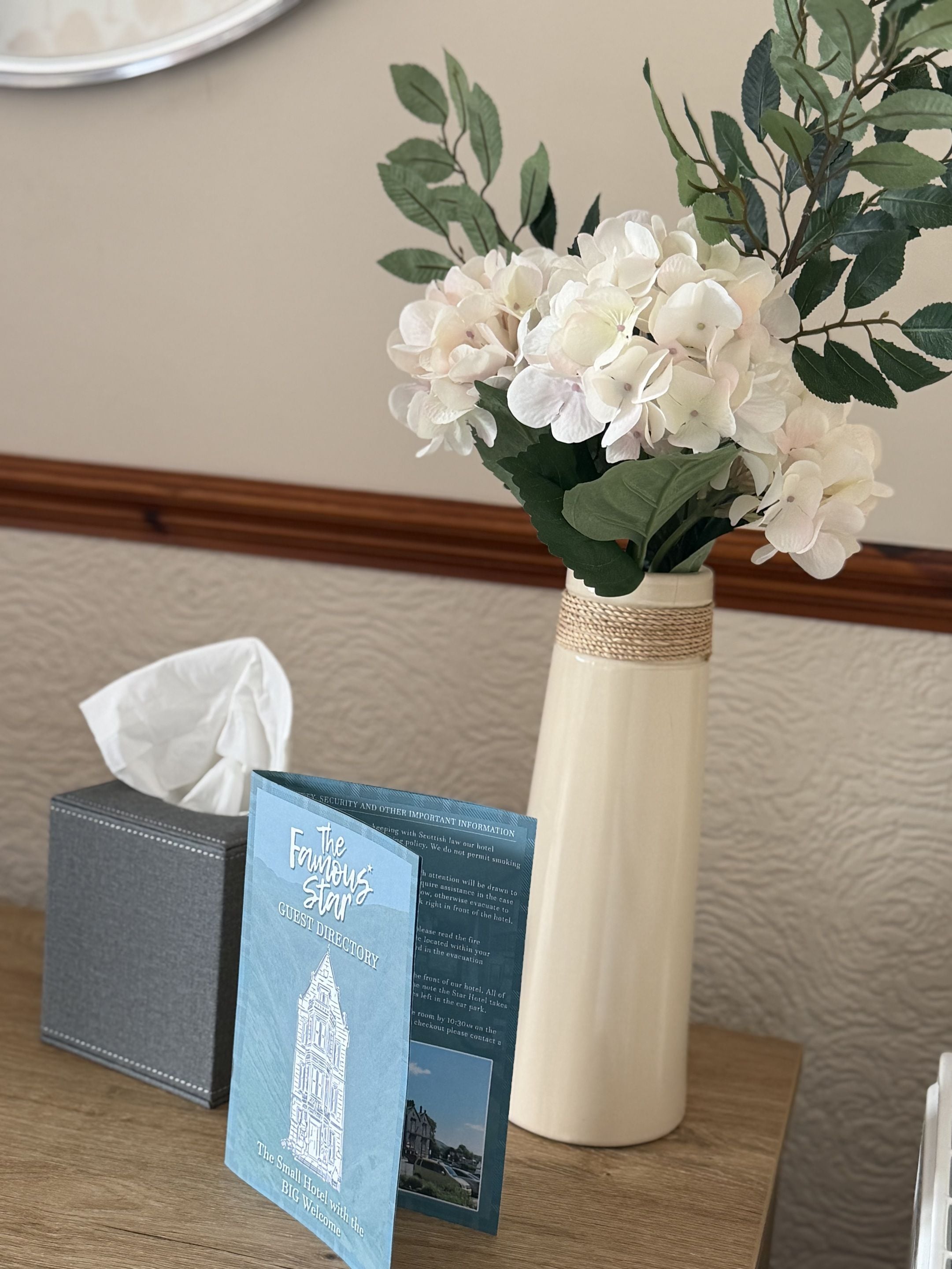 Una mesa con un jarrón de flores blancas, una caja de pañuelos y un directorio para huéspedes impreso.