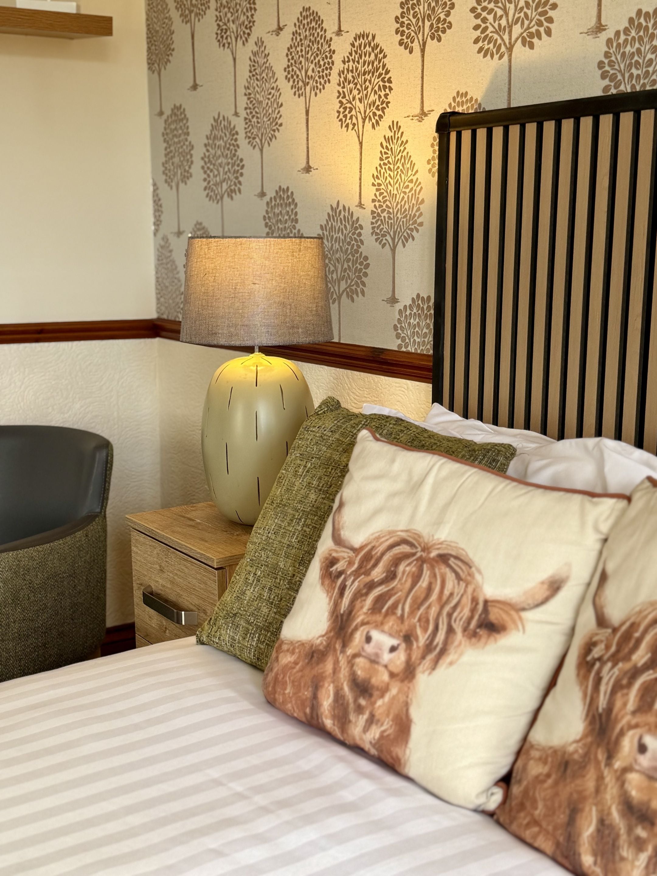 A hotel room with a bed, a nightstand with a lamp, a chair, and a wall with a tree pattern.