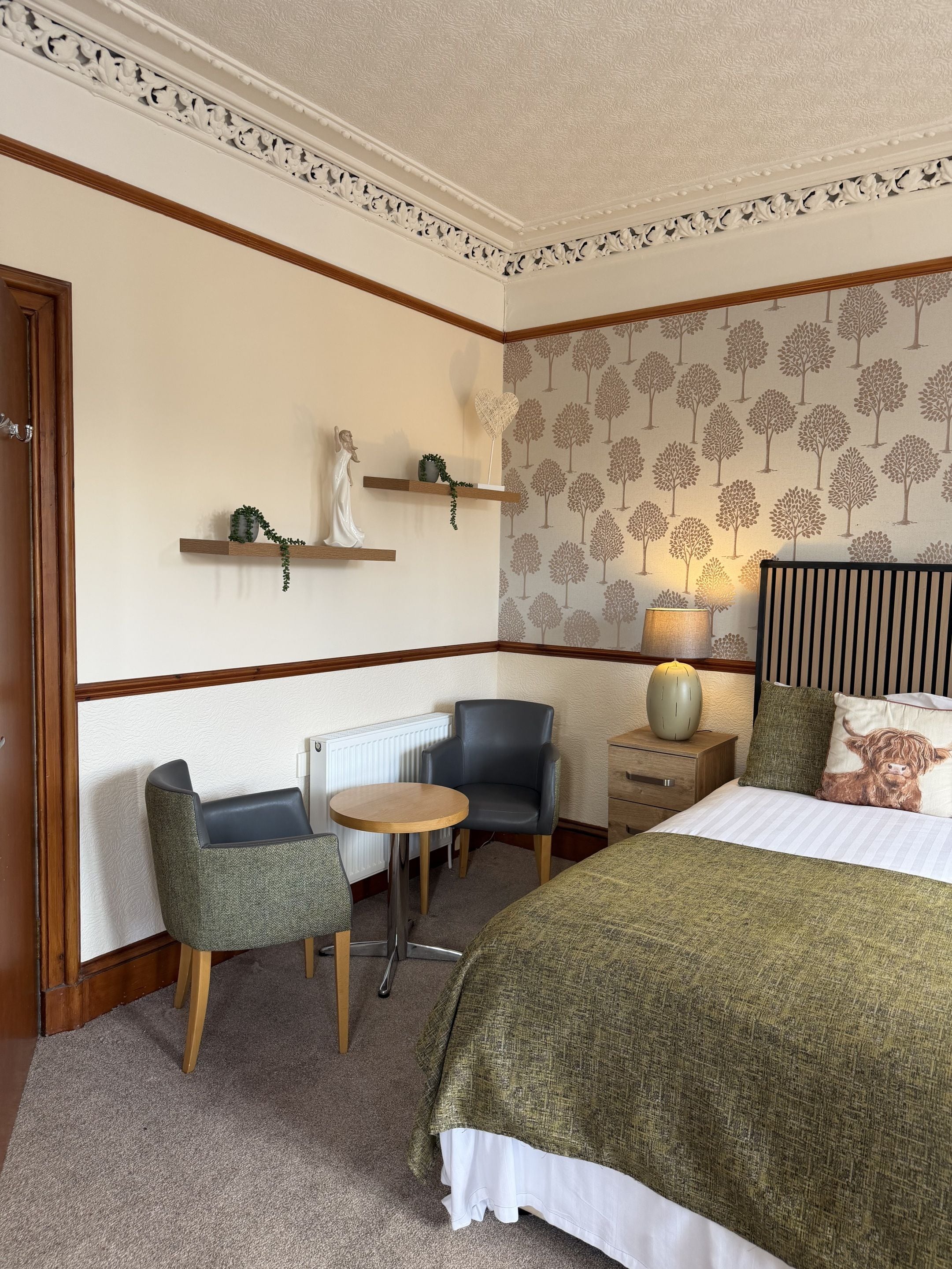 A hotel room with a bed, a chair, a small table, a nightstand, and a wall with a tree pattern.