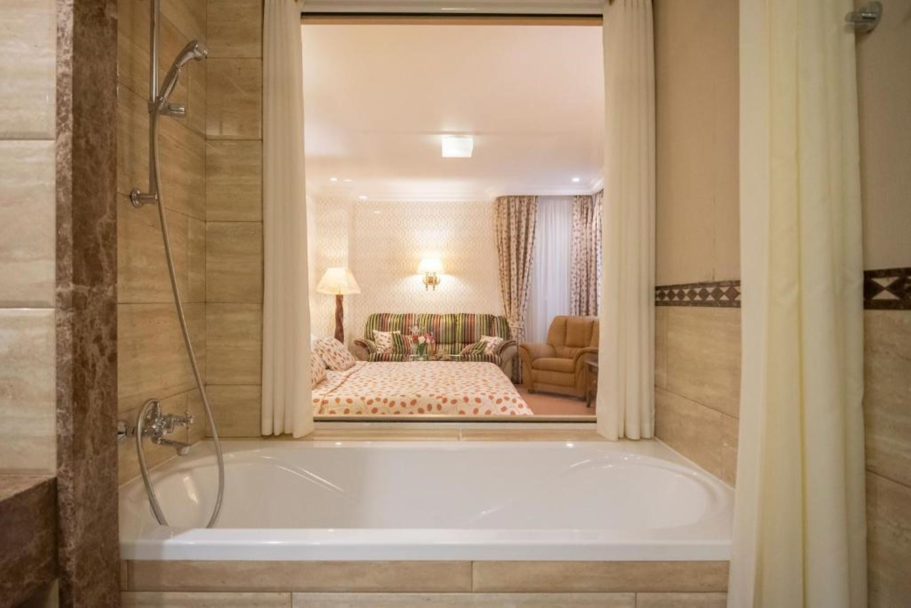 A bathroom with a bathtub, shower, and a view into a bedroom with a bed, a chair, and a lamp.