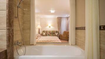 A bathroom with a bathtub, shower, and a view into a bedroom with a bed, a chair, and a lamp.