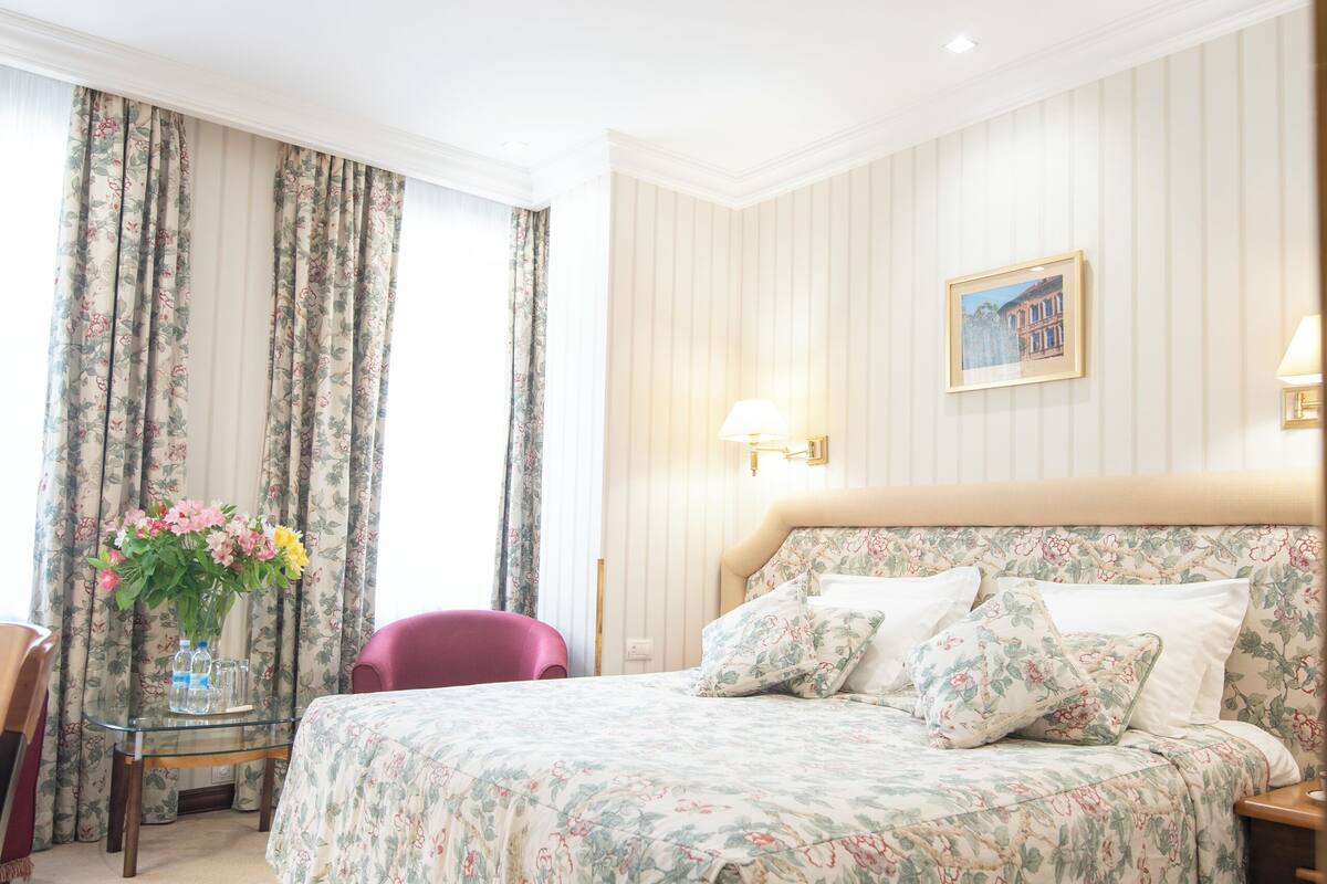 A hotel room with a large bed, floral bedding, a small table with a vase of flowers, a chair, and a picture on the wall.