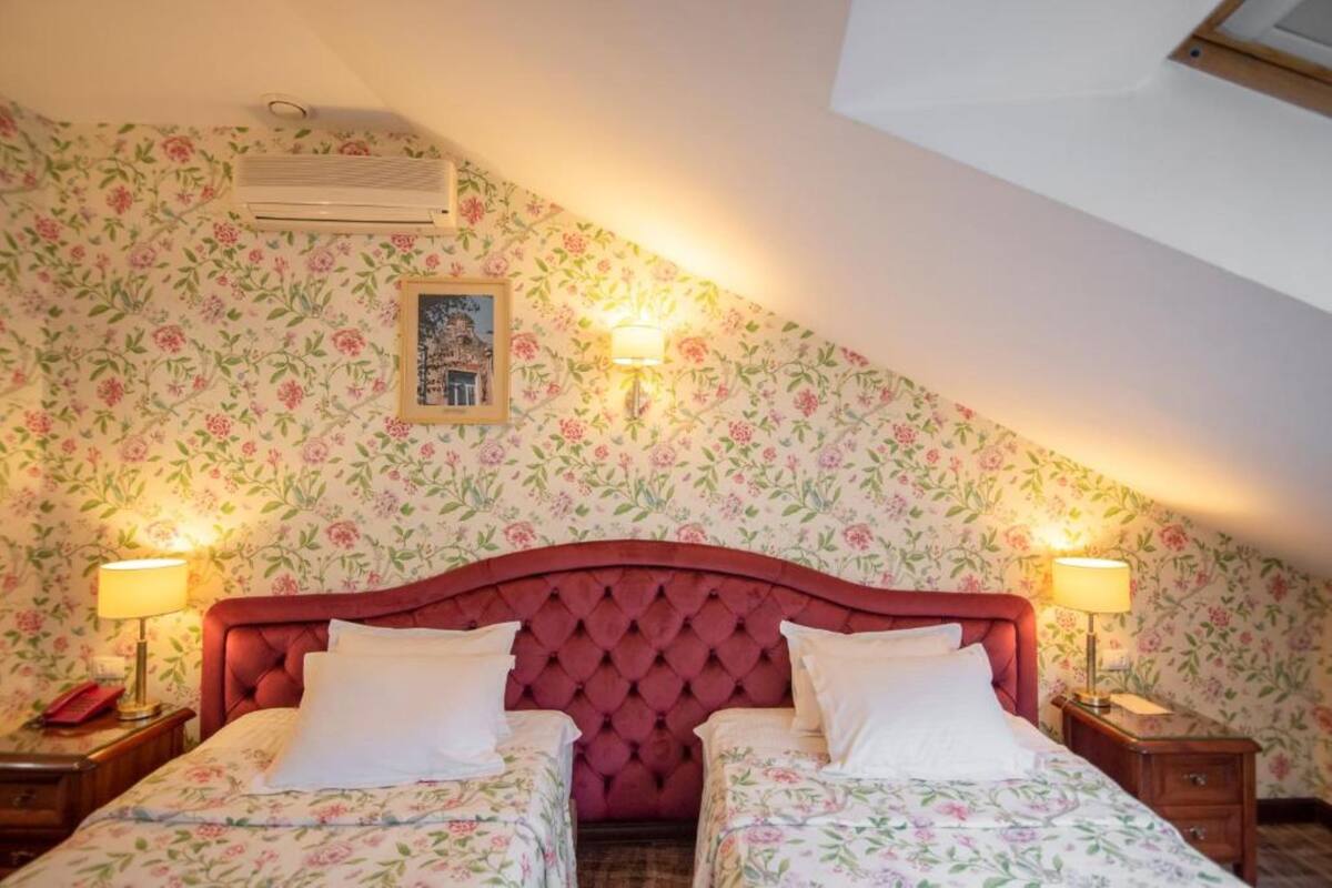 A bedroom with two beds, floral wallpaper, a red headboard, bedside lamps, and an air conditioning unit.