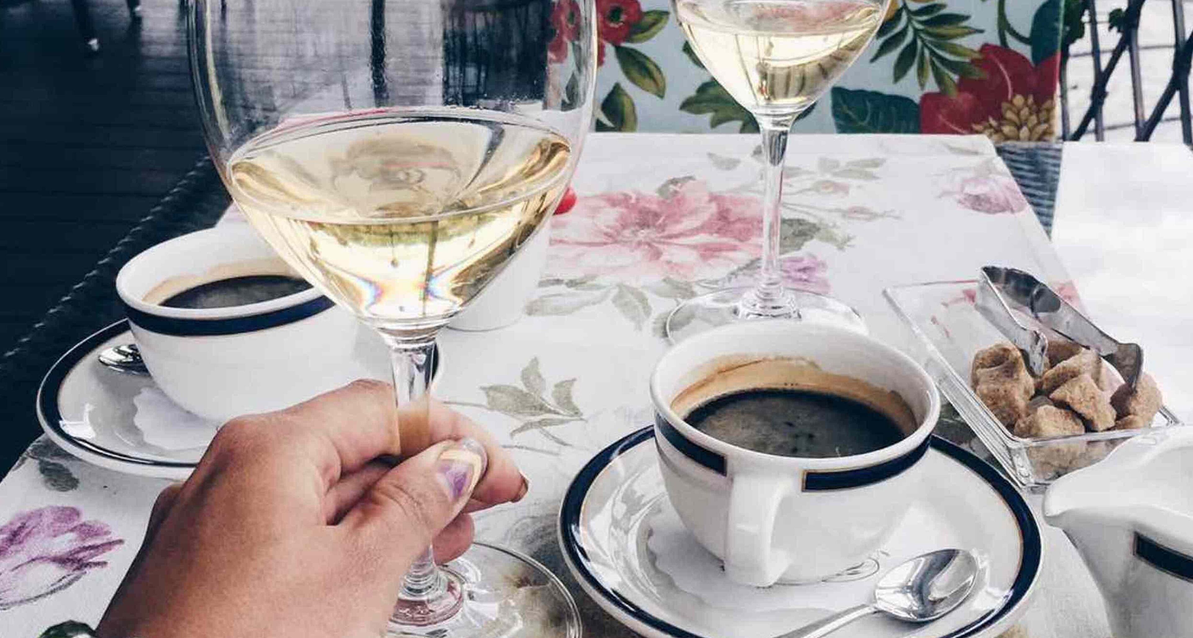 A table with a floral tablecloth, a glass of white wine, two cups of coffee, and a small container of biscotti.