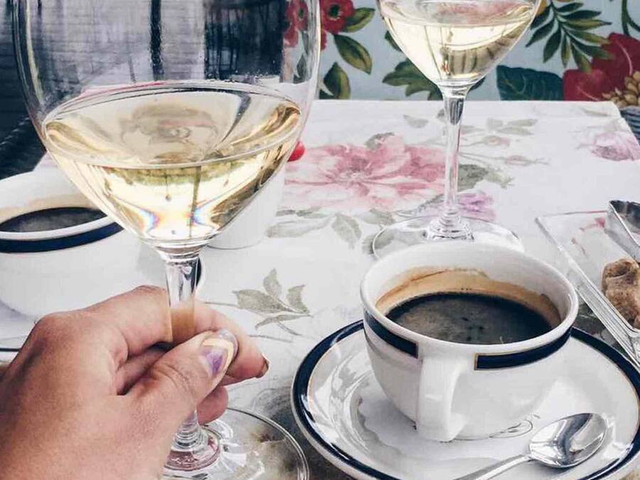 A table with a floral tablecloth, a glass of white wine, two cups of coffee, and a small container of biscotti.