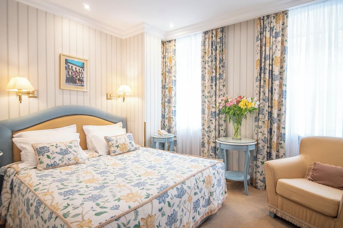 A hotel room with a large bed, two bedside lamps, a floral-patterned bedspread, a small round table with a vase of flowers, a sofa, and curtains with a floral design.
