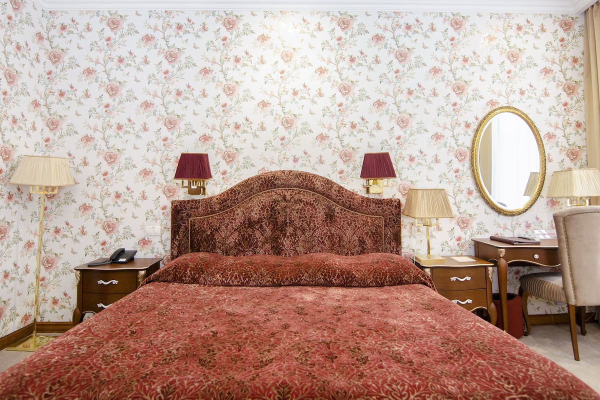 A bedroom with a floral wallpaper, a wooden headboard, two bedside tables with lamps, a chair, and a mirror.