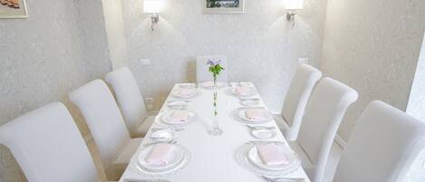 Un comedor con una mesa larga dispuesta para una comida, sillas blancas e iluminación empotrada en la pared.