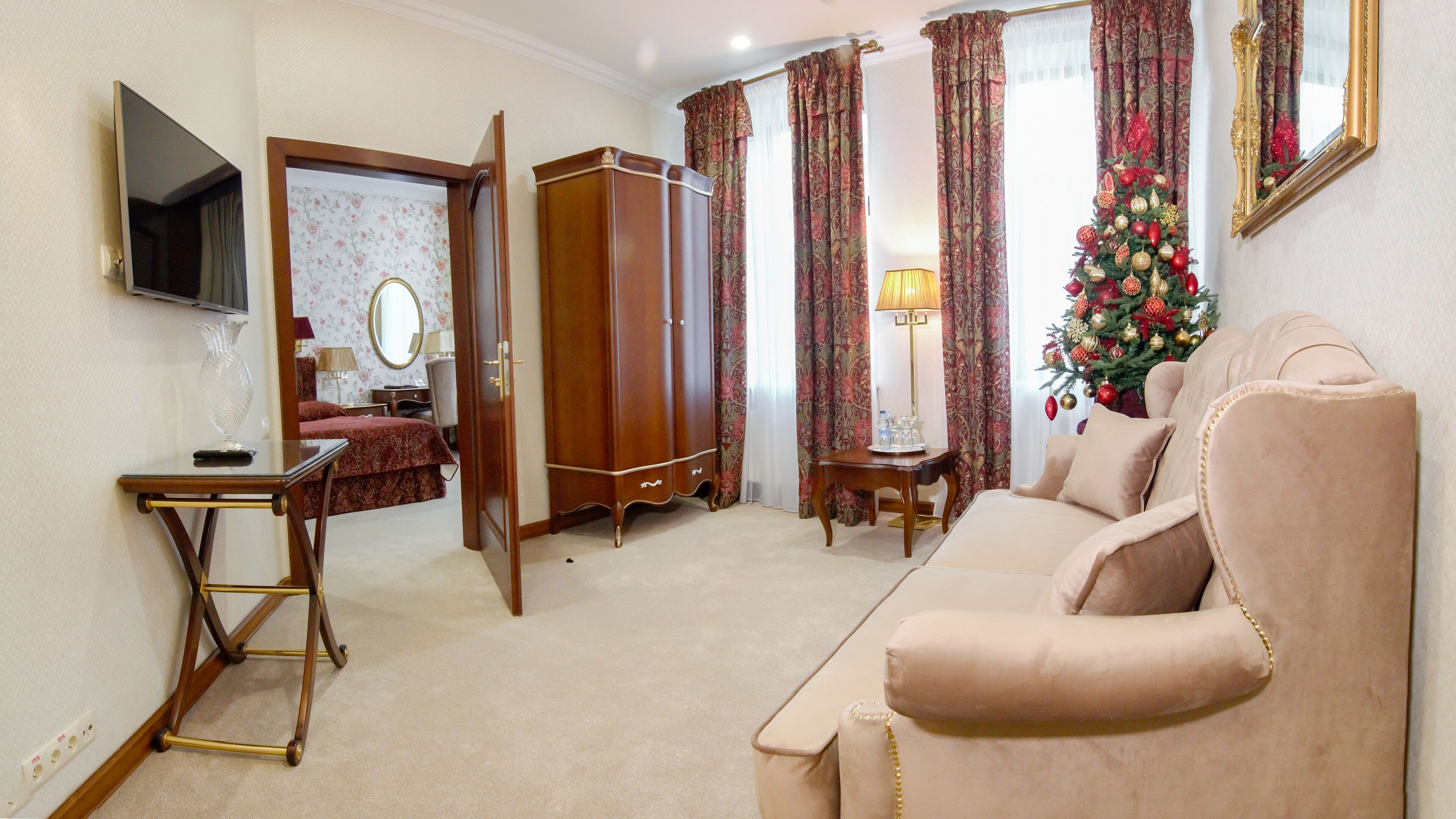 Une pièce bien éclairée, avec un canapé beige, une armoire en bois, un bureau avec une lampe et un sapin de Noël.