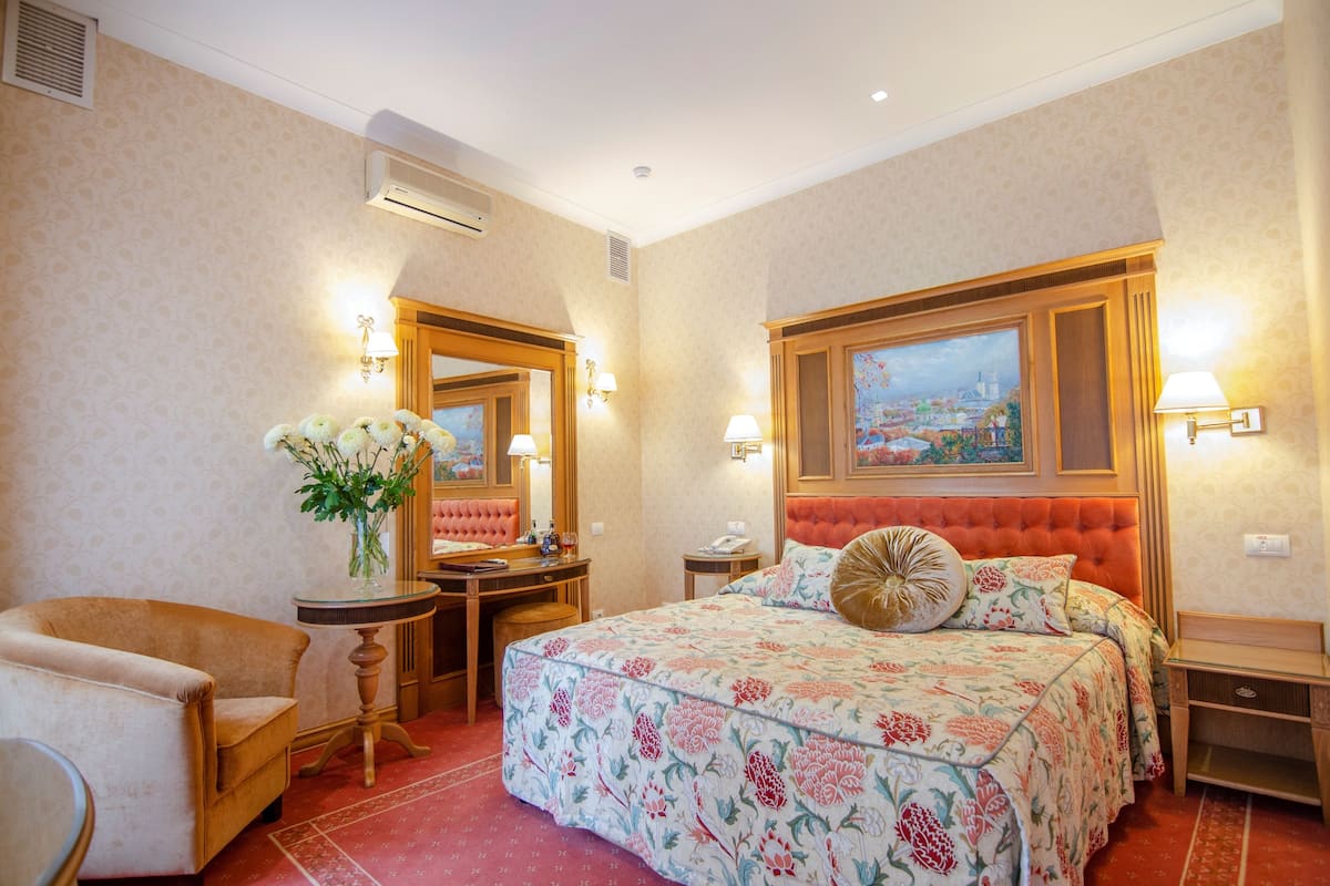 A hotel room with a large bed, a desk with a chair, a small table with a vase of flowers, and a nightstand.