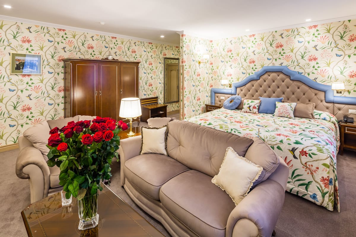 A bedroom with a floral wallpaper, a tufted headboard, a sofa, a bed with floral bedding, and a vase of red roses on a coffee table.