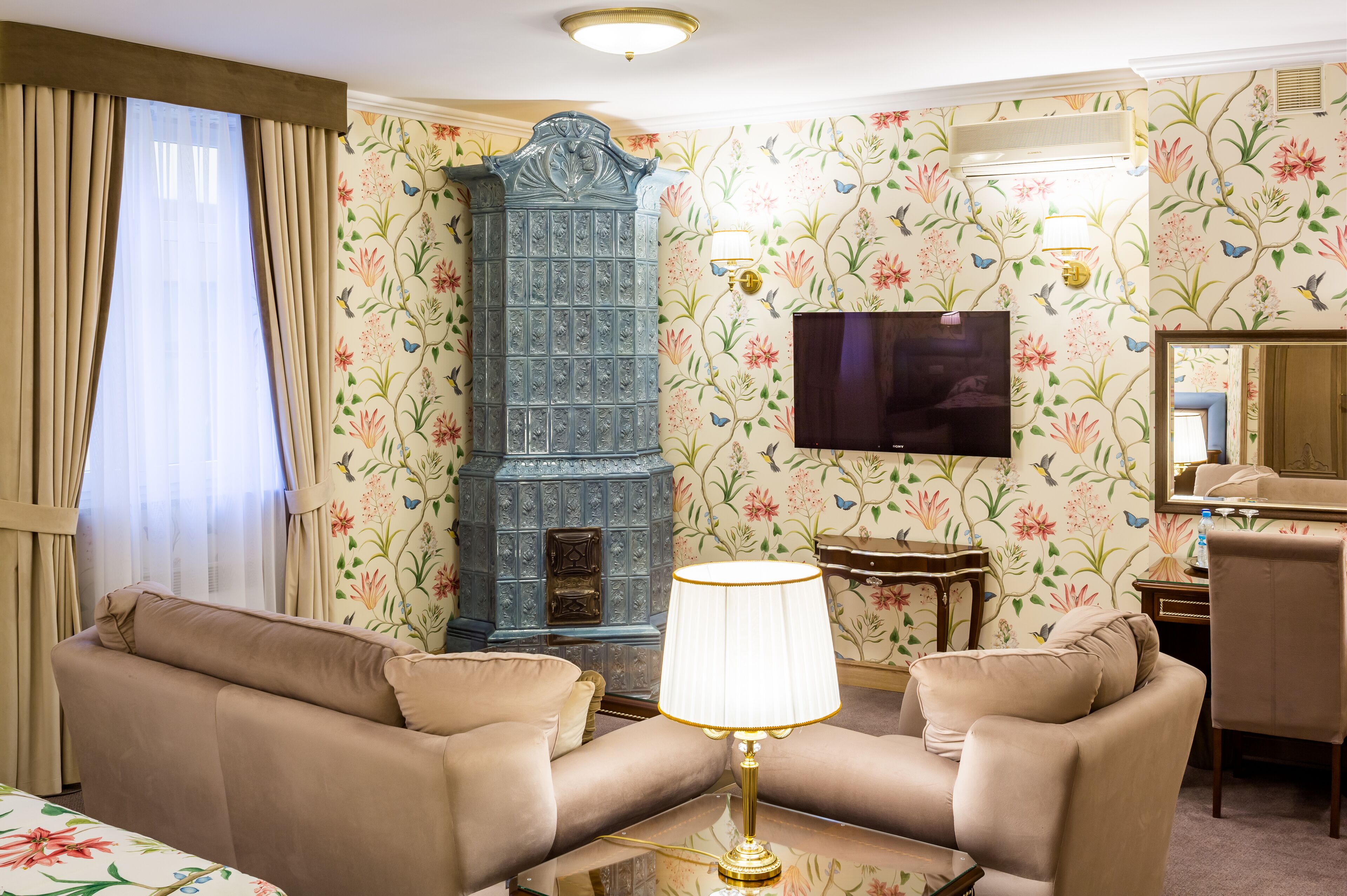 A living room with beige sofas, a table with a lamp, a blue cabinet, and floral wallpaper.