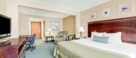 A hotel room with a large bed, a desk with a TV, a chair, a lamp, and framed artwork on the wall.