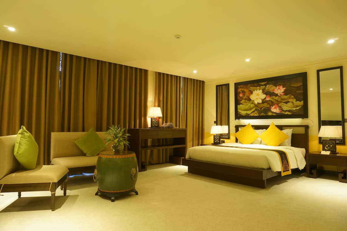 Habitación de hotel con cama, sofá, mesita de noche, lámpara, un cuadro y un ventanal grande.