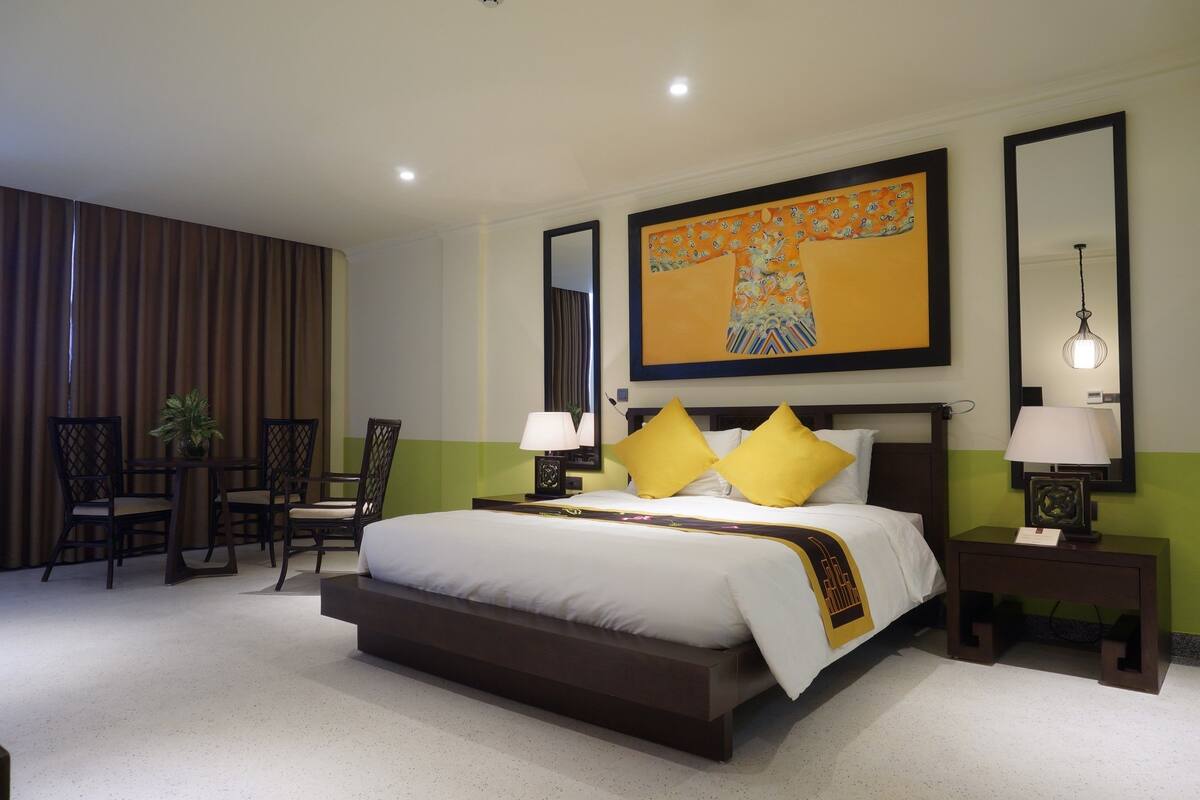Un cuarto de hotel con una cama grande, dos mesitas de noche con lámparas, una silla, una pequeña mesa con una planta y un cuadro grande en la pared.