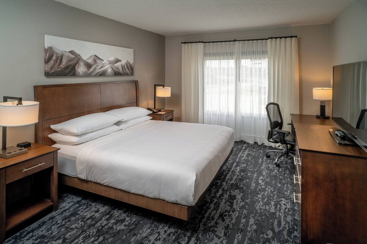 A hotel room with a large bed, two bedside tables with lamps, a desk with a chair, and a window with curtains.