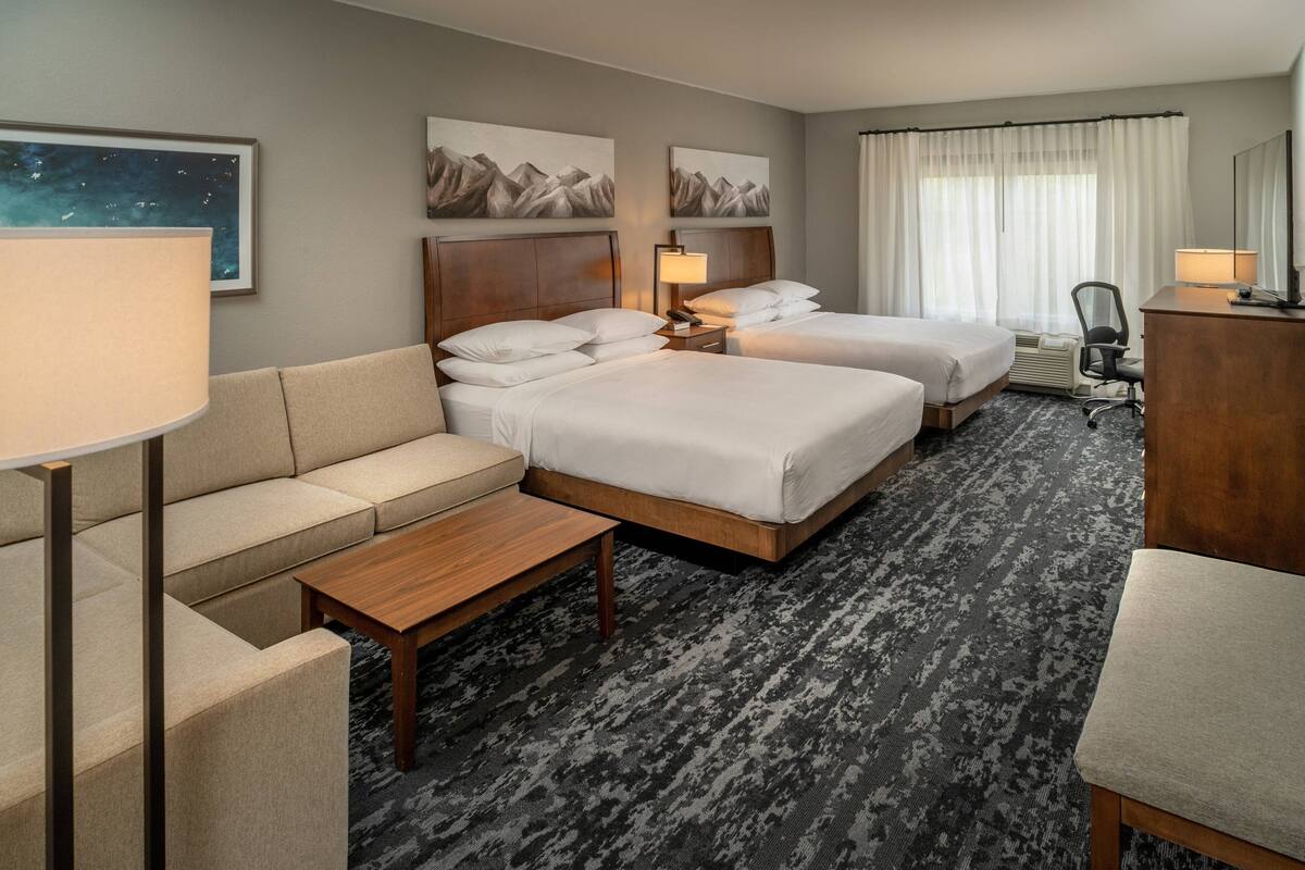 A hotel room with two beds, a sofa, a desk, and a chair. There are paintings on the wall and a lamp on the desk.