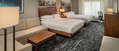 A hotel room with two beds, a sofa, a desk, and a chair. There are paintings on the wall and a lamp on the desk.