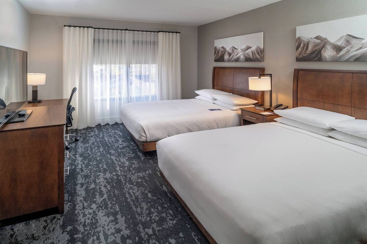 A hotel room with two beds, a desk, a chair, a lamp, and a painting of mountains.