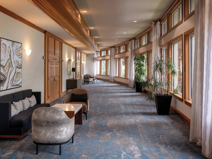 Un vestíbulo de hotel con un largo pasillo, zona de estar con sofás y sillones, ventanas amplias y plantas en macetas.