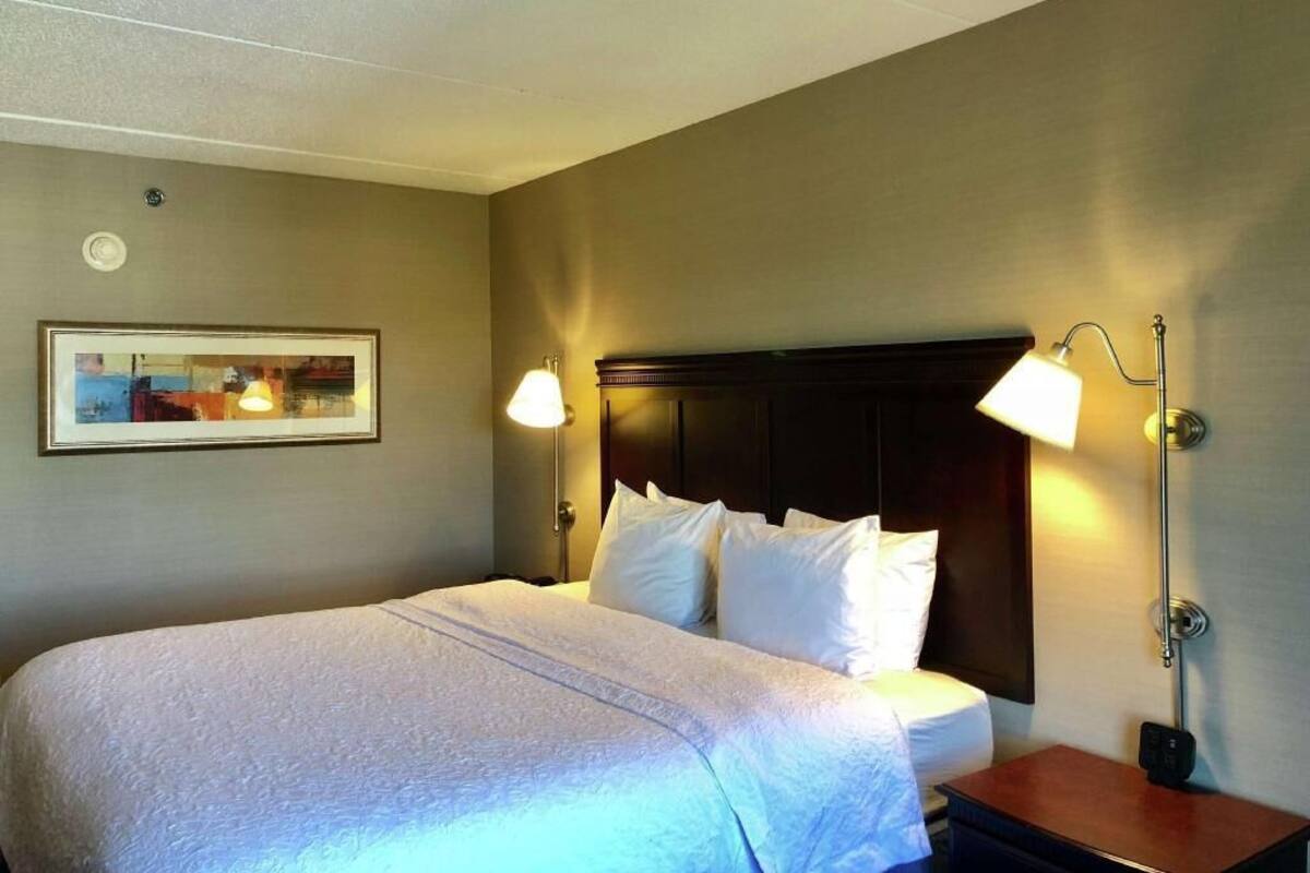 A hotel room with a bed, two bedside lamps, a framed picture on the wall, and a nightstand.