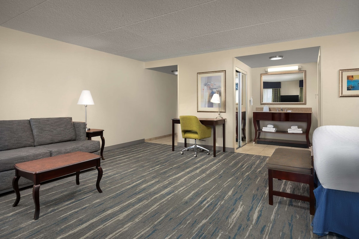 A hotel room with a sofa, a desk, a chair, a small table, a bed, a bathroom with a mirror, and a framed picture on the wall.