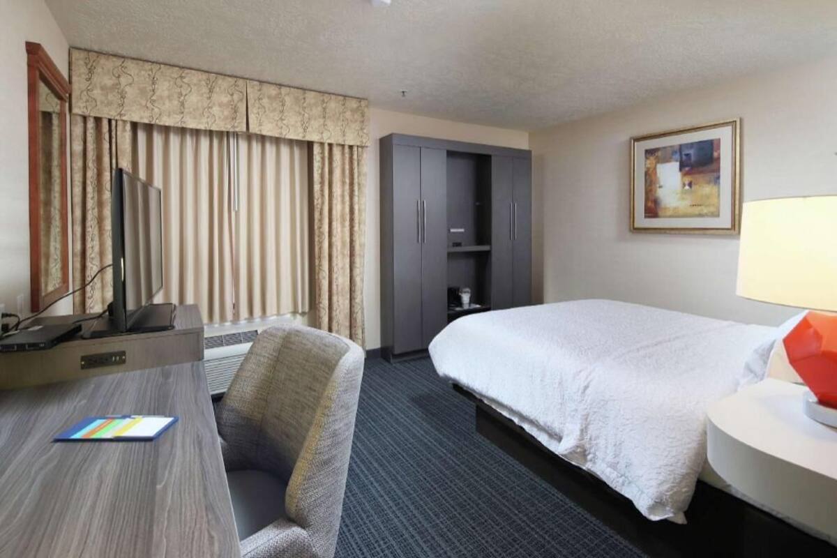 A hotel room with a bed, a desk with a chair, a wardrobe, and a painting on the wall.