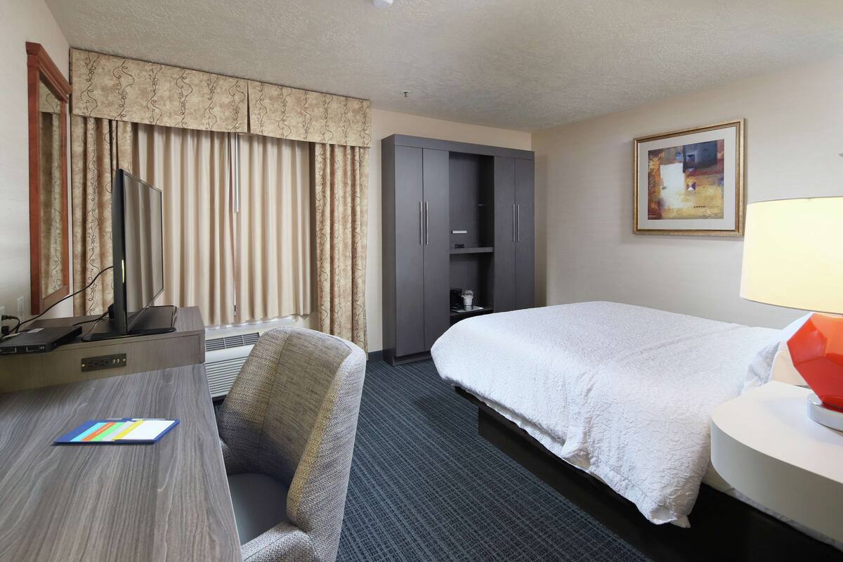 A hotel room with a bed, a desk with a chair, a wardrobe, a painting on the wall, and a lamp.