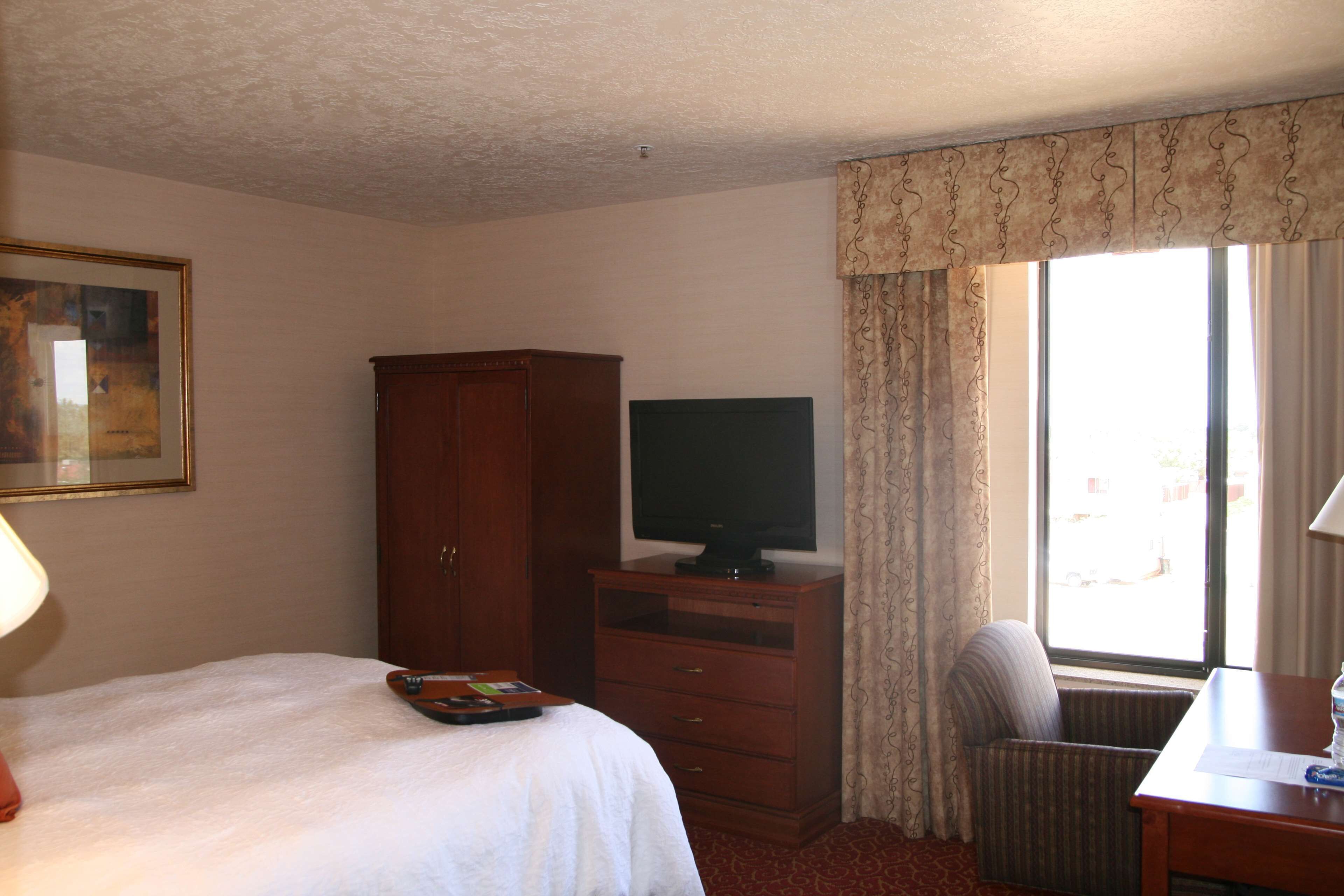 Quarto de hotel com uma cama, um armário de madeira, uma televisão, uma cadeira e uma escrivaninha com abajur.