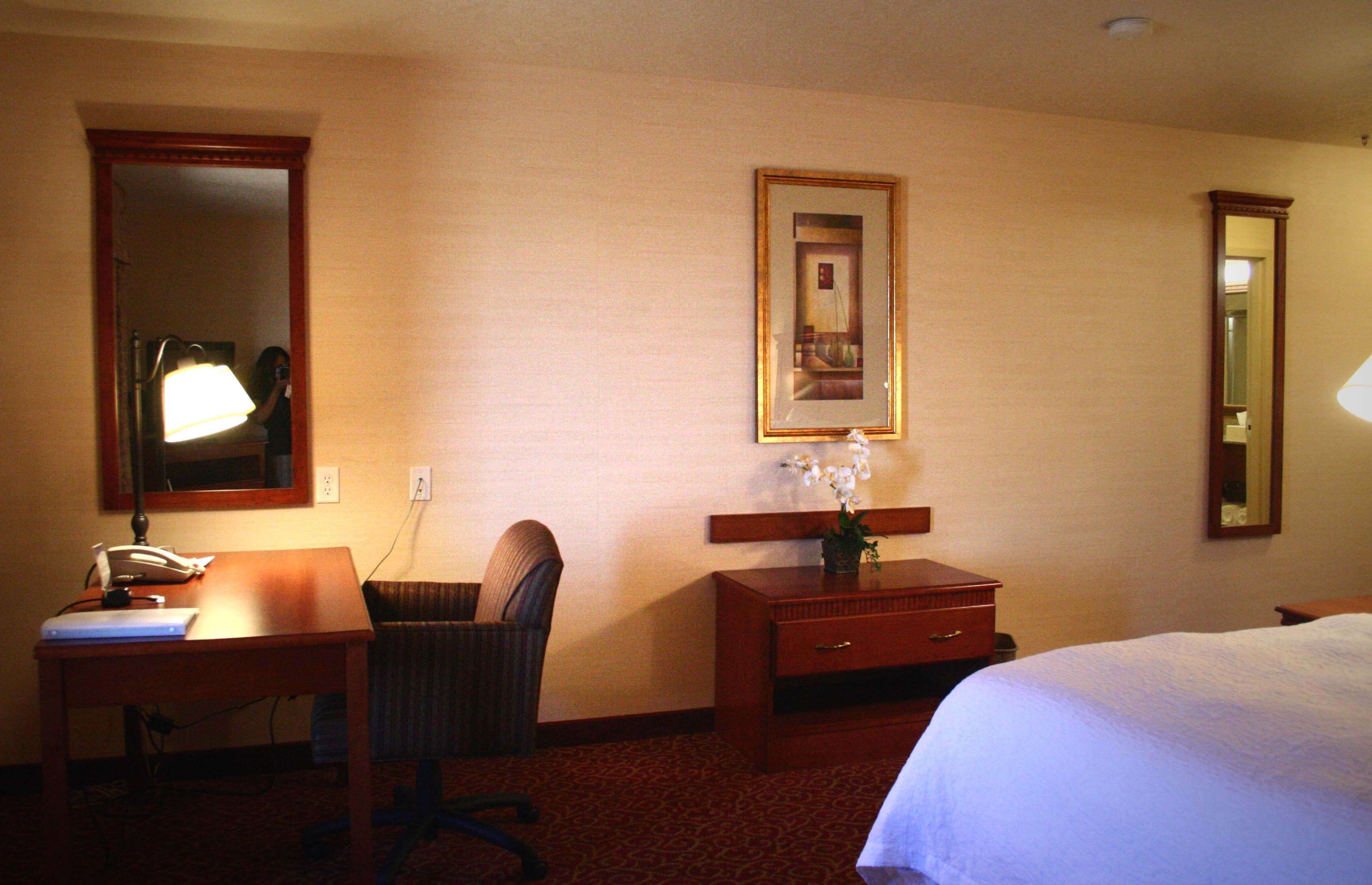 A hotel room with a bed, a desk with a lamp, a chair, a dresser, and a mirror.