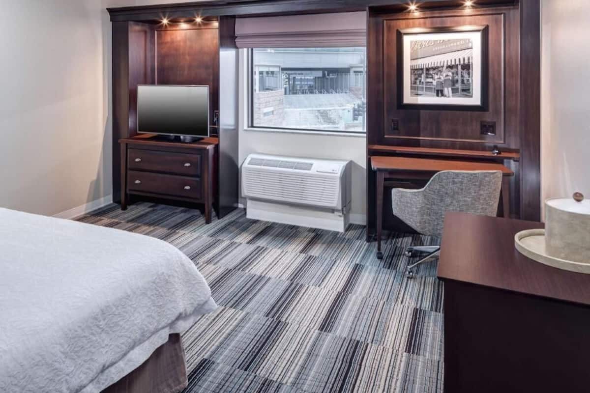 A hotel room with a bed, a desk with a chair, a flat-screen TV, a window with a view of a building, and a framed picture on the wall.