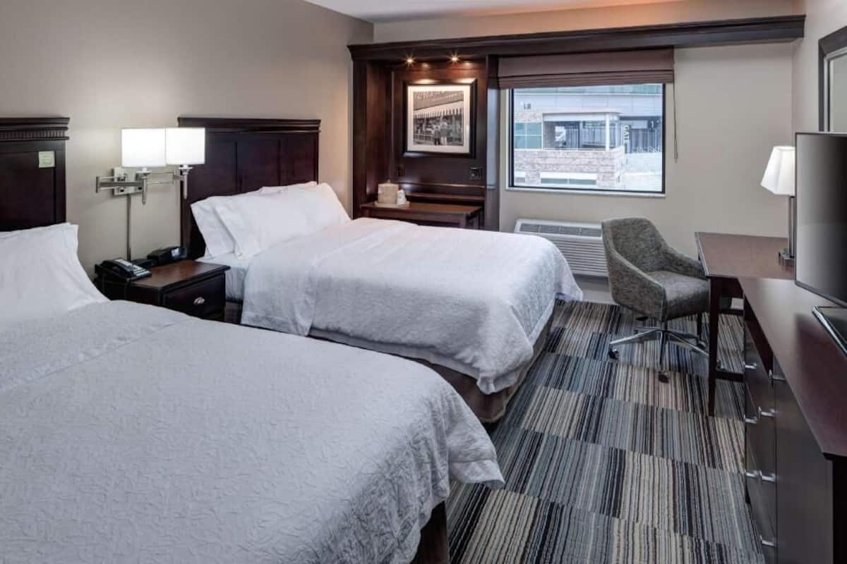 Quarto de hotel com duas camas, uma escrivaninha, uma cadeira, uma televisão, uma janela com vista e uma luminária.