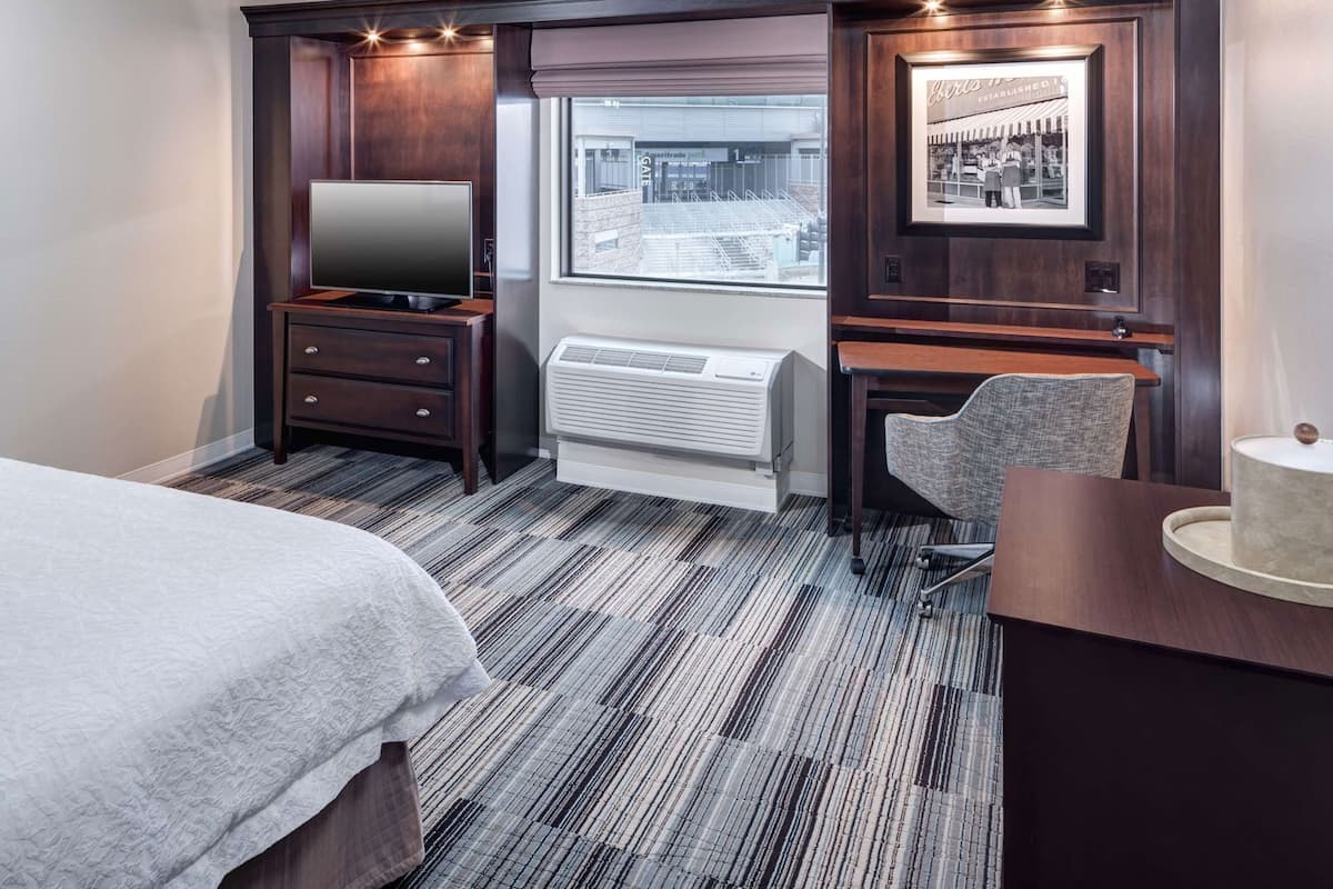 Quarto de hotel com cama, escrivaninha, cadeira, televisão, aquecedor e janela com vista para um edifício.