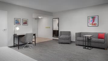 A modern room with a grey carpet, a grey sofa with a red pillow, a grey armchair, a small table, and a desk with a chair. There are two framed pictures on the wall.