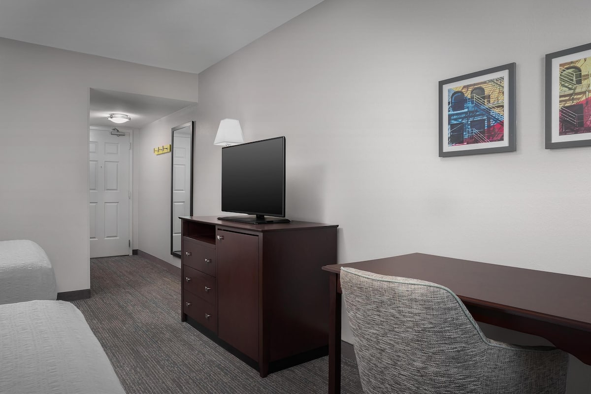 A hotel room with a bed, a desk, a chair, a TV on a dresser, and two framed artworks on the wall.