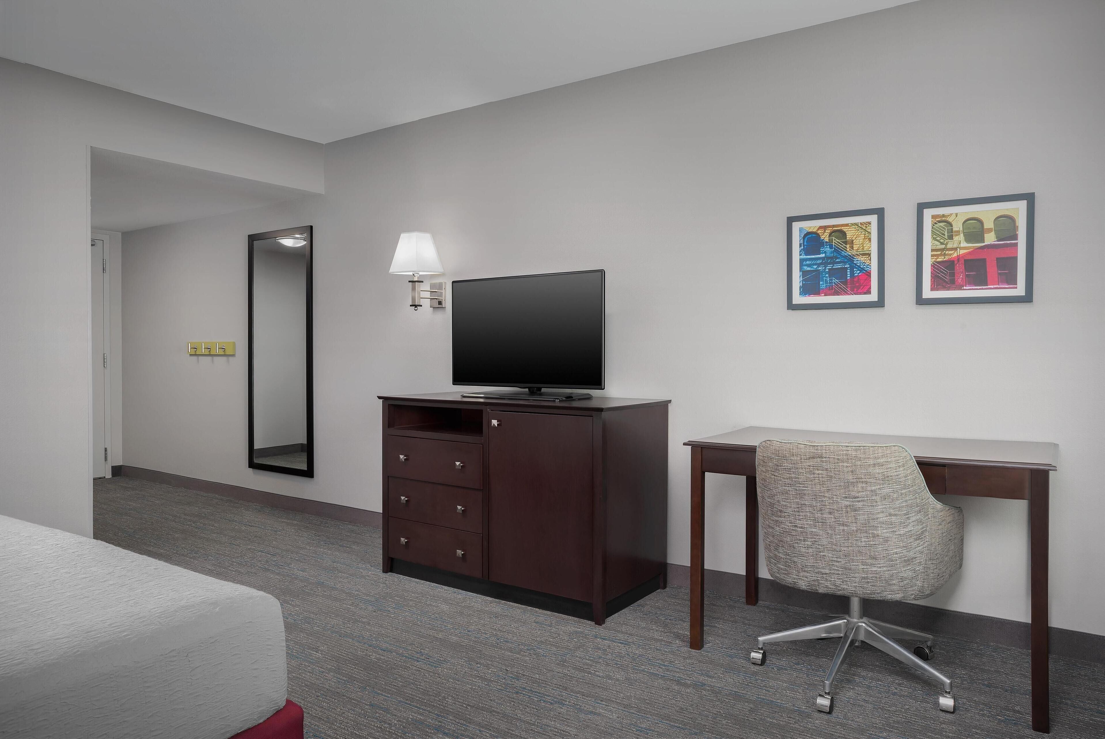 A hotel room with a bed, a desk with a chair, a TV, a dresser, and two framed pictures on the wall.